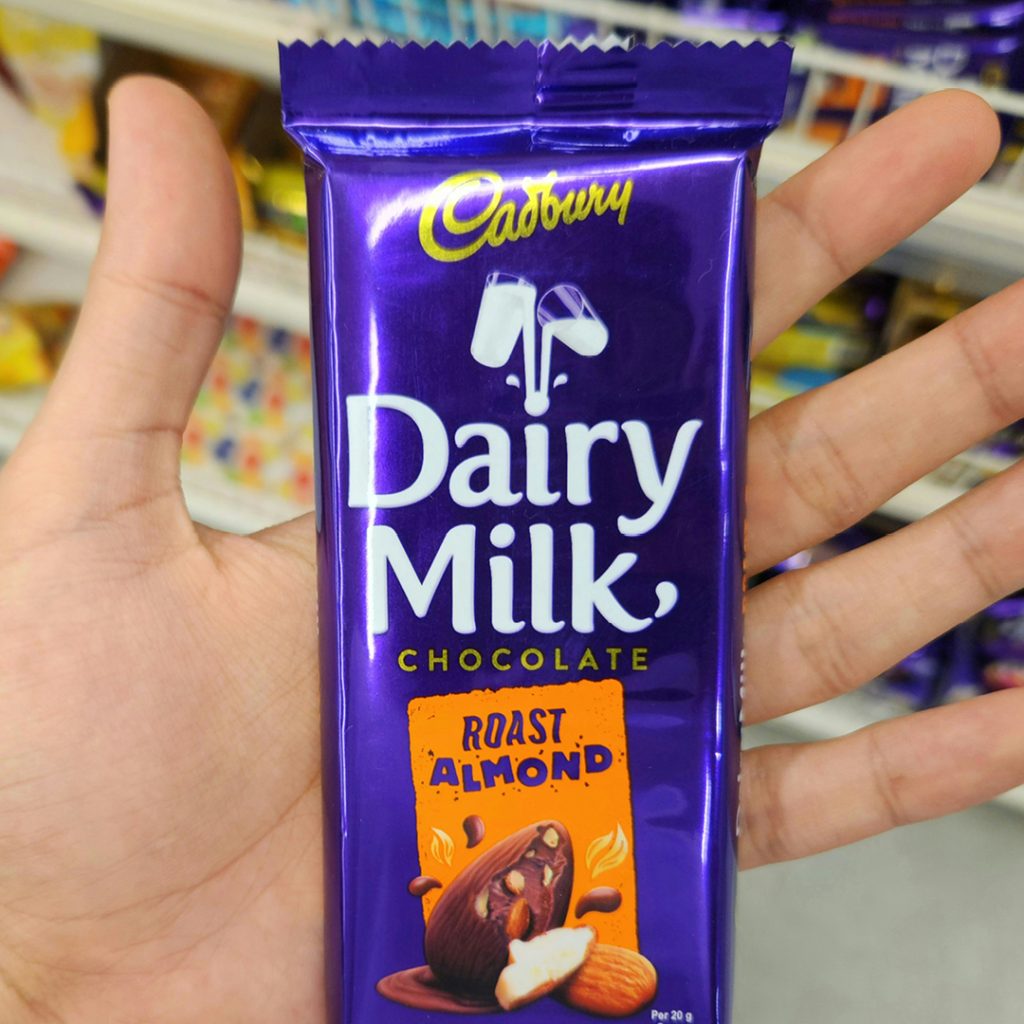 A hand holding a Cadbury Dairy Milk Roast Almond chocolate bar in a store aisle.